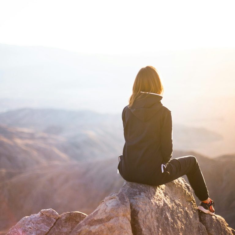 Eine Person sitzt auf einem Felsen und blickt über eine bergige Landschaft im Sonnenlicht.